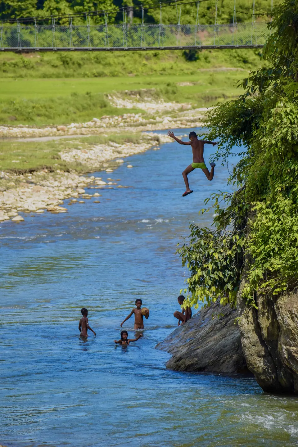 People crossing a clear river below a wooded hillside.